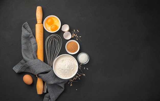 Flour, eggs, sugar and rolling pin on a black background. Culinary background, baking. Top view, copy space.