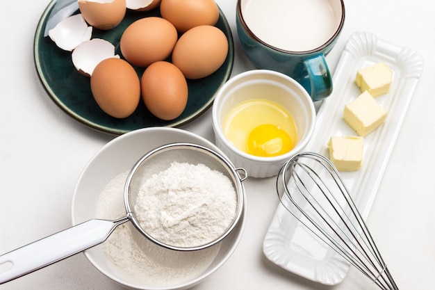 Flour in sieve and in bowl. Butter and whisk on plate. Egg yolk in bowl. Milk in blue mug. Brown eggs on blue plate. White background. Top view