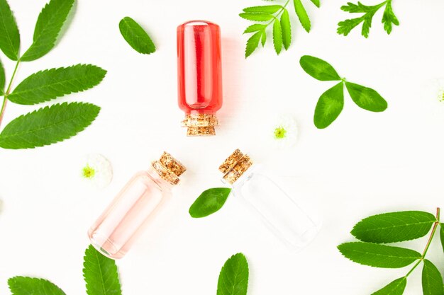 Flower extracts in glass bottles on a white wooden background with green leaves and flowers top view