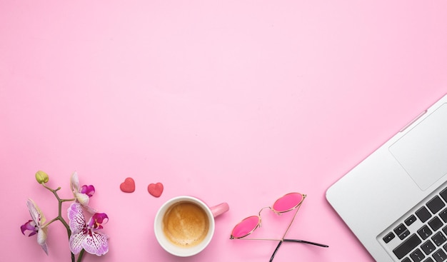 Flowers cup of coffee and laptop on pink desk background copy space