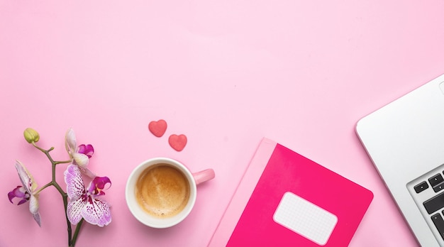 Flowers cup of coffee and laptop on pink desk background copy space