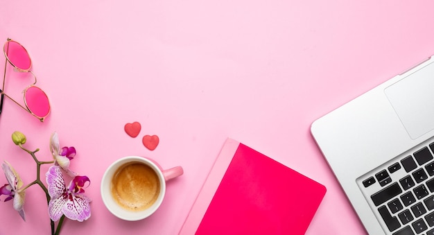 Flowers cup of coffee and laptop on pink desk background copy space