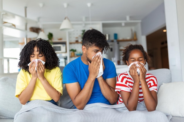 Flu And Illness Concept Portrait of sick young African American family blowing noses with napkins together while sitting on the couch covered with blanket Four unwell people with cold at home