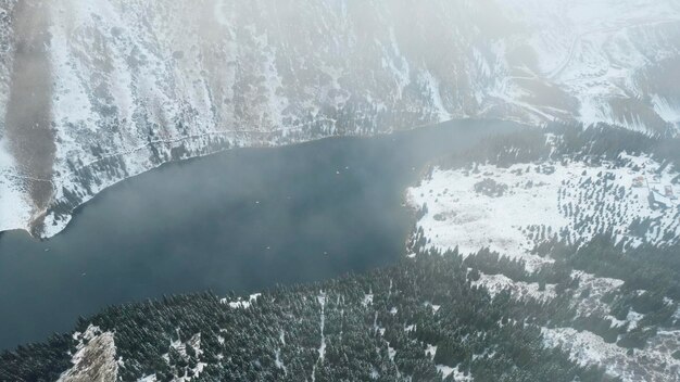 Flying in the clouds over the mountain lake Kolsai. Top view of snowy mountains and forest.