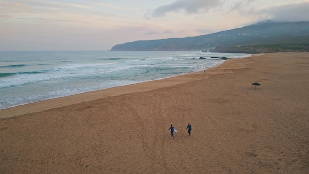 Foamy sea waves rolling on sandy beach drone view surfers walking seashore