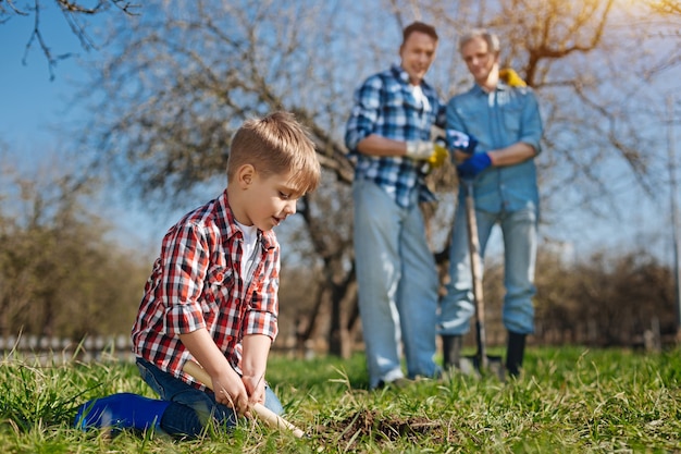 Focus on a little boy standing on knees while digging into the soil with a stainless scoop with his dad and grandfather behind looking at him