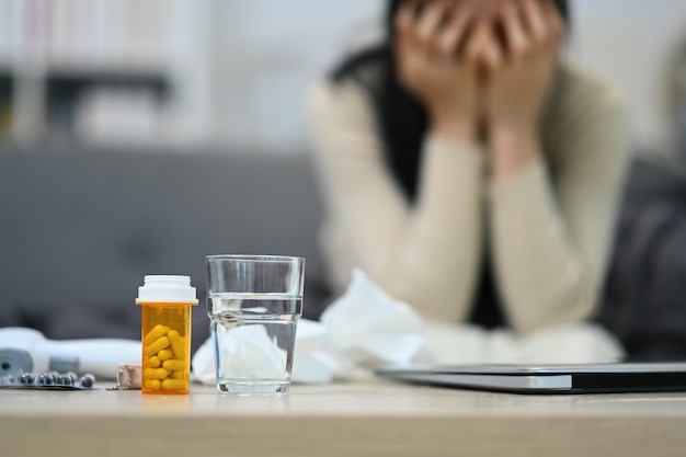 Focus on pills bottles and glass of water with sick woman sitting in background Medicine pharmacology healthcare concept