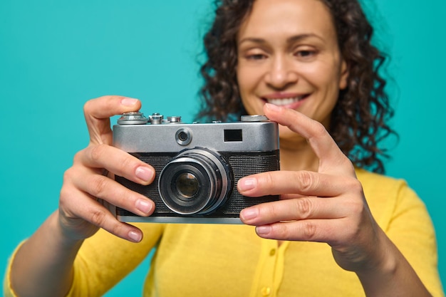 Focus on a vintage camera- old retro style photographic equipment in the hands of a blurred smiling cheerful woman with curly hair, dressed in bright yellow sweater, isolated over blue background.