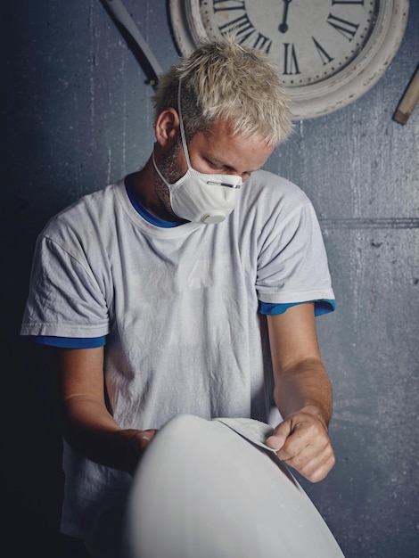 Focused adult blond haired tattooed male master grinding white handmade surfboard while shaping it with sandpaper in workshop