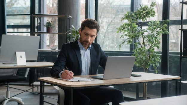 Focused adult businessman work on project in office using laptop writing notes caucasian man