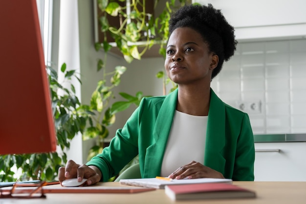 Focused African American woman sits at computer desk holding mouse in hand and looking at monitor