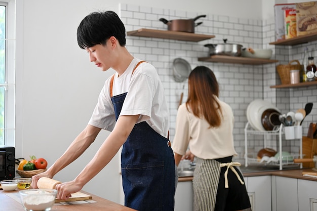 Focused Asian man is making dough while his girlfriend is washing the dishes in the kitchen