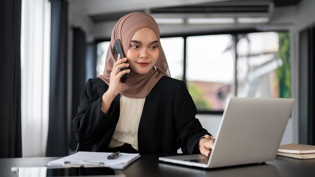 A focused Asian Muslim businesswoman is talking on the phone while checking details on her laptop