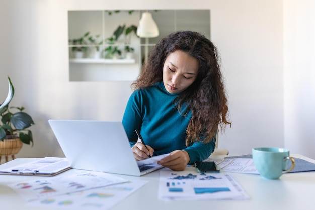 Focused Asian woman using laptop at home looking at screen chatting reading or writing email sitting on couch female student doing homework working on research project online