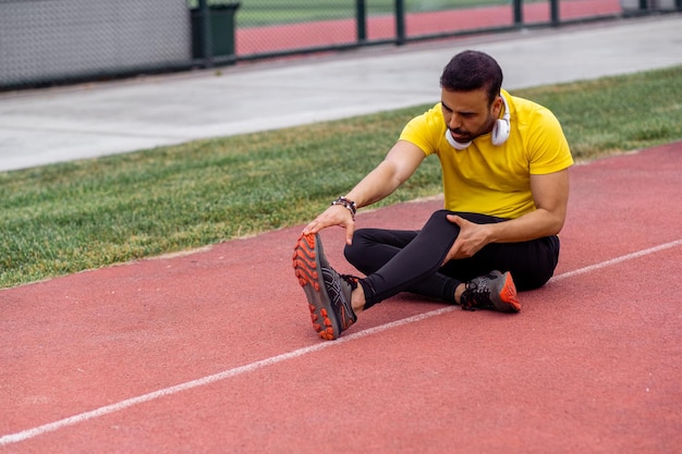 Focused athlete wearing earphones performs butterfly stretch on stadium track in a vibrant city park