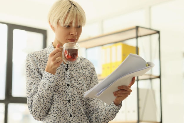 Focused blonde woman drinks aromatic tea reading accounting papers female employee with short