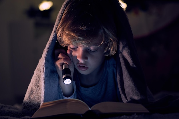 Focused boy reading book with interest and shining flashlight while lying on bed under warm blanket in dark room of apartment