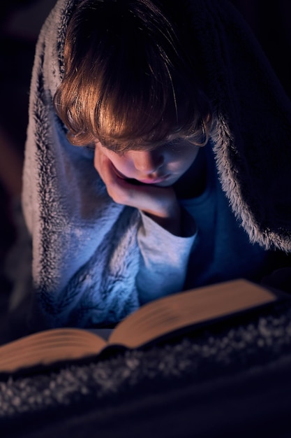 Focused boy wrapped in warm blanket reading on hand and reading interesting book while lying on bed in dark room