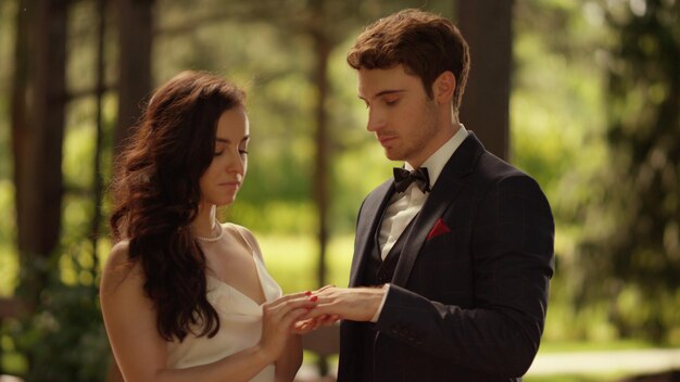 Focused bride putting ring on groom finger on wedding ceremony in park Newlyweds man and woman getting married outdoors Closeup wedding couple standing in garden