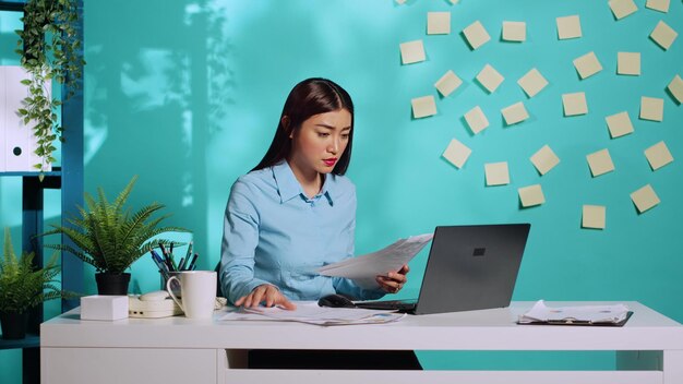 Focused businesswoman executive cross-referencing invoicing administrative data between paperwork and laptop. Professional office clerk working in modern workplace over blue studio background