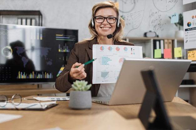 Focused caucasian senior woman in headset sitting at desk with paper report in her hands