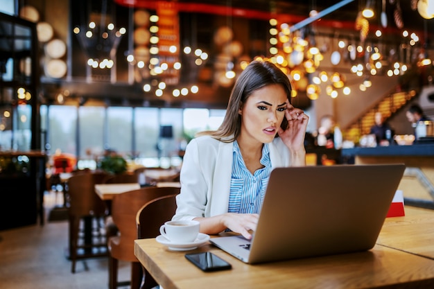 Focused charming fashionable businesswoman sitting in cafe and using laptop for work.