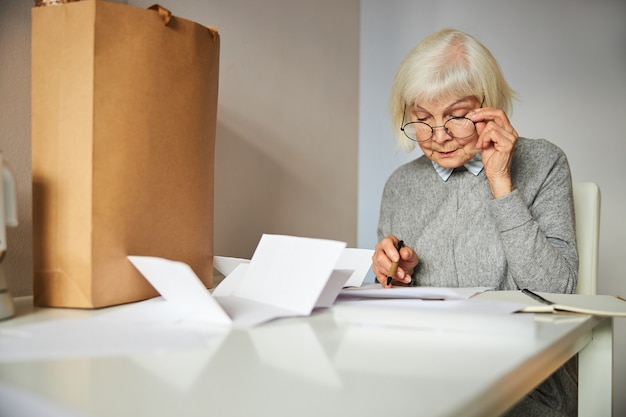 Focused elderly blonde Caucasian lady in eyeglasses staring at the utility bills on the table