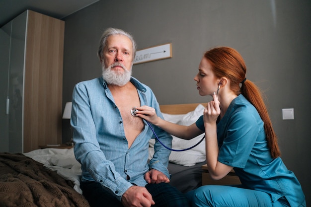 Focused female nurse holding stethoscope listening lungs old senior male patient sitting on bed at