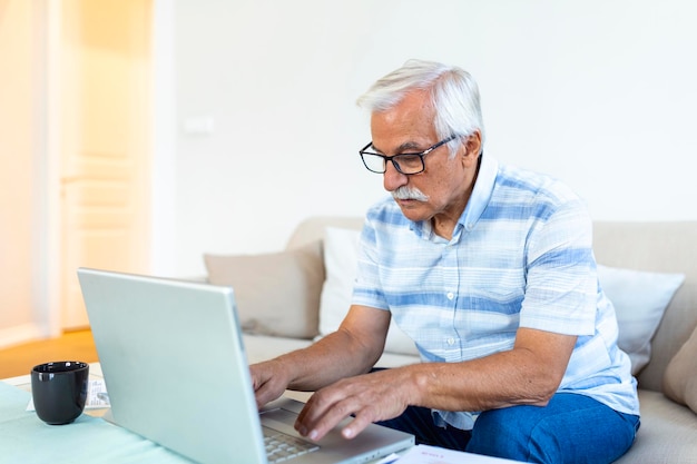 Focused greyhaired elderly man sit on couch reading bank notifications calculating domestic expenditures concentrated modern mature male consider financial paperwork pay bills on laptop online