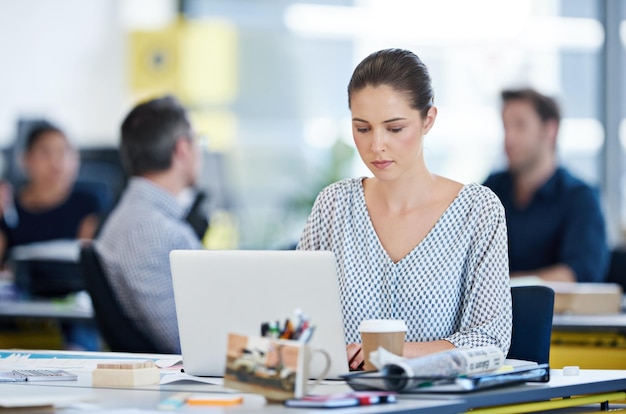 Focused on her tasks Shot of a designer sitting at her desk working on a laptop with colleagues in the background