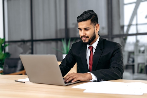 Focused intelligent confident arabian or indian successful businessman company ceo in formal suit sit at work desk with laptop in modern creative office concentrated looks at laptop screen