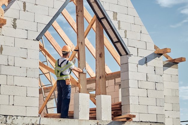 Focused male builder wearing safety gear while standing on top of the house and putting a nail in the wood.