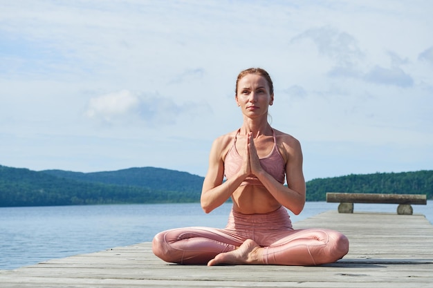 Focused mature woman in a pink tracksuit practices yoga on a wooden bridge near the sea