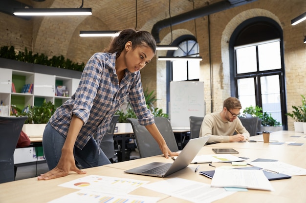 Focused mixed race young business woman standing in the modern coworking space looking at laptop