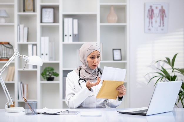 A focused muslim female doctor in a hijab and white coat examines documents in her medical office