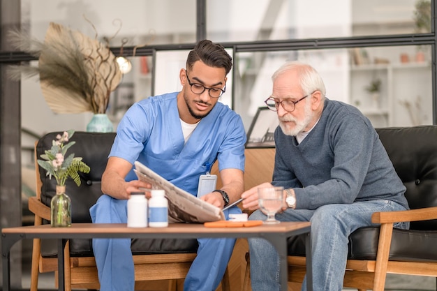 Focused old man seated at the coffee table with his caregiver reading an article in the newspaper