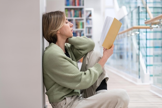 Focused pensive middleaged woman passionate reader reading book while sitting on floor in library