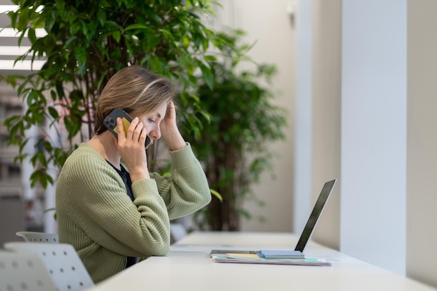 Focused pensive woman talking on mobile phone while working remotely in library reading room