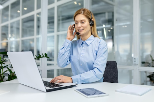 A focused professional woman in a blue shirt working with headphones at a bright office workspace