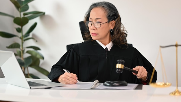 Focused senior female lawyer in robe gown uniform sitting front of laptop providing law consultation and legal advice online