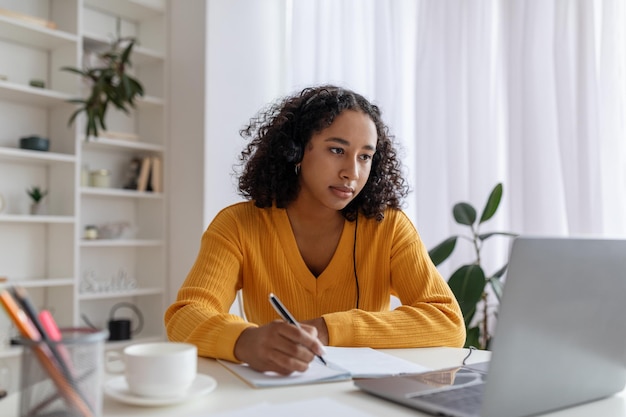 Focused young black woman in headset having online lesson on laptop writing in notebook at home