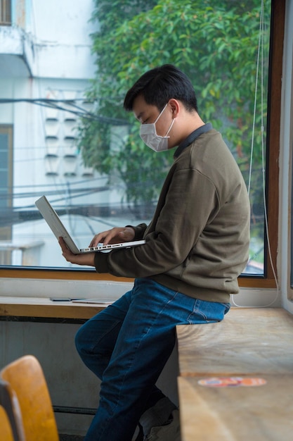 Focused young man wearing a medical mask using laptop typing on keyboard writing email