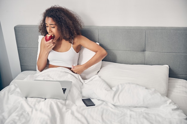 Focused young woman biting off a piece of apple in front of her laptop