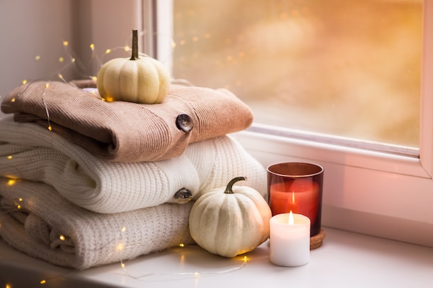 Folded knitted sweaters pumpkins and candles near the window Autumn concept