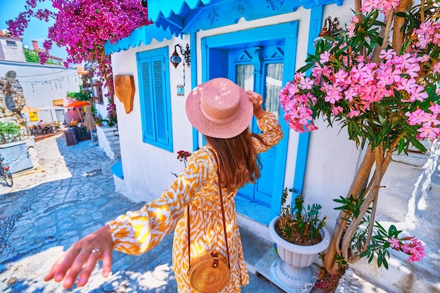 Follow me concept Girl traveler wearing dress and hat walks on beautiful colorful flower street with white houses and blue doors in a European city