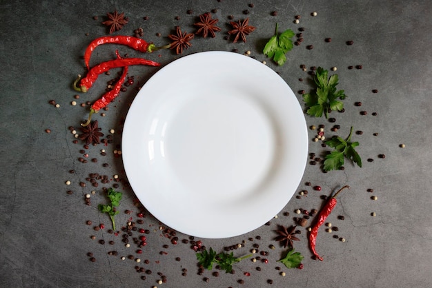 Food background An empty plate with space for text among seasonings and spices Chili pepper allspice star anise and greens on a dark background