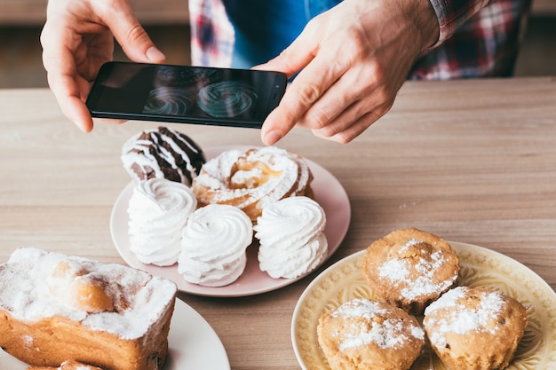 Food blogger. Dessert. Mobile photography. Man with smartphone taking photos of fresh homemade pastries. Meringue, fruitcake and muffins.