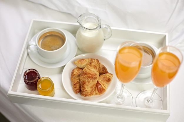 food, breakfast and eating concept - close up of tray with croissants, coffee, milk and two glasses of orange juice in bed