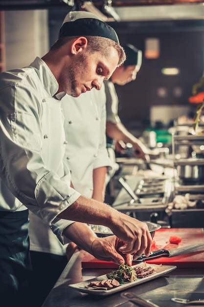 Food concept young serious chef in white uniform decorate beef steak meat dish with green herbs and