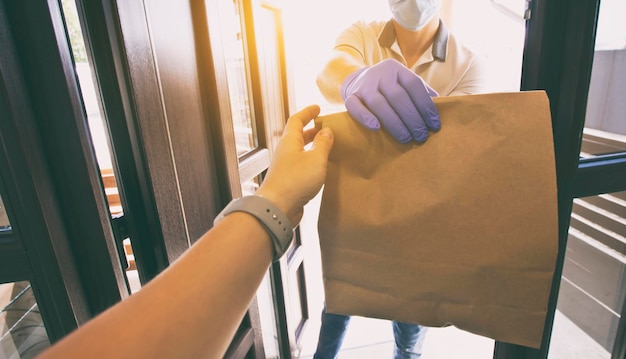 The food delivery guy wearing gloves and face mask is giving the order to the customer at home